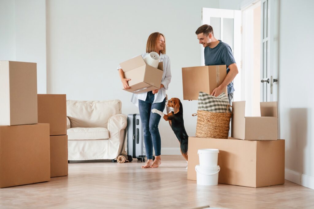 An image of a man and a woman and their dog holding boxes and walking into their new home.